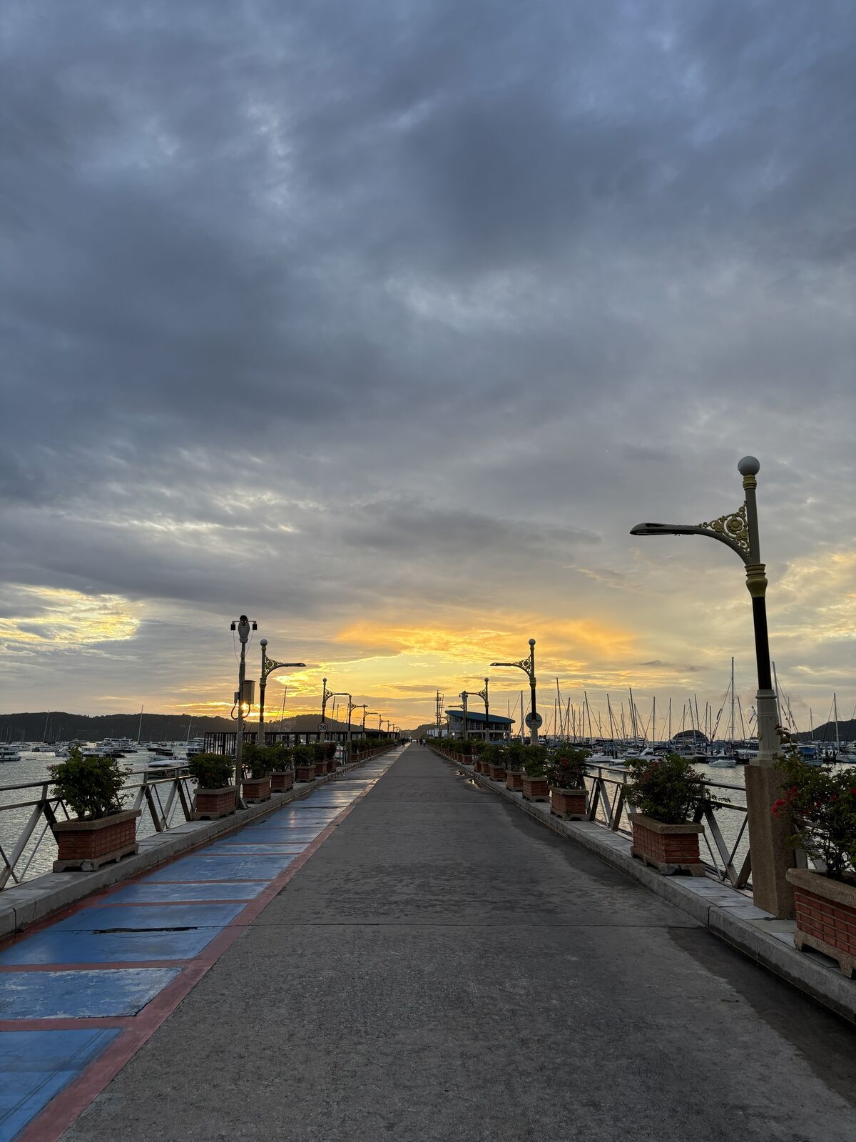 Chalong Pier at dusk: the long walkway with lamp posts and planters leading out to moored boats under a streaked sky