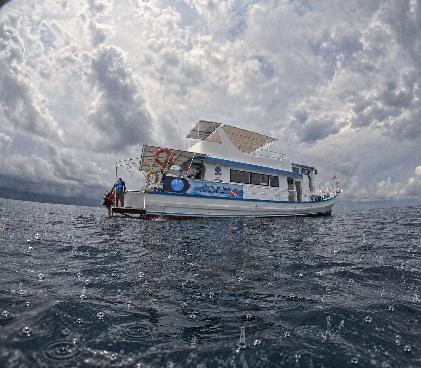MV Matchanu at anchor under a heavy grey sky, raindrops dimpling the sea in the foreground