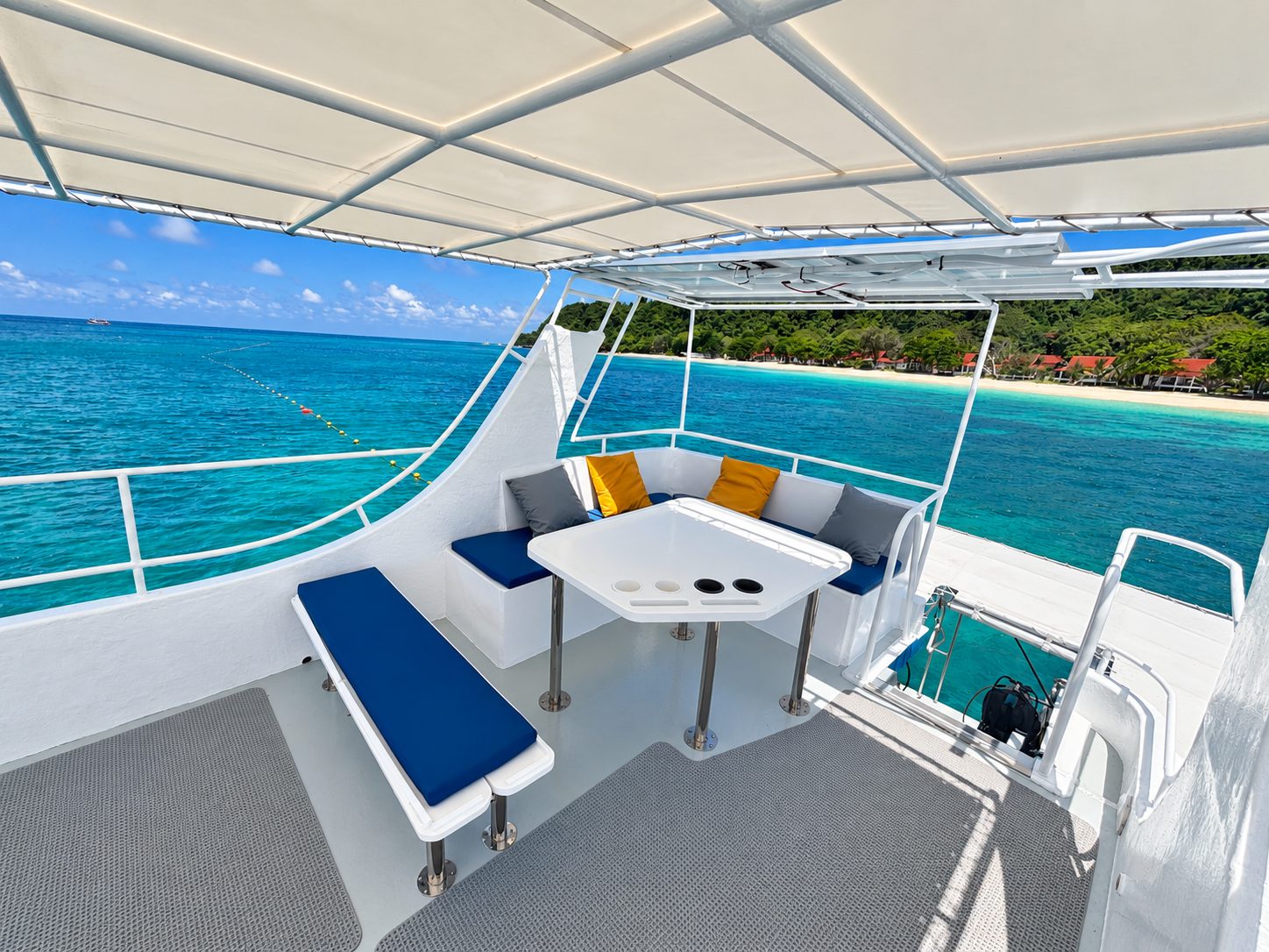 The shaded sun deck: blue cushioned benches around a table, a white beach and palms beyond