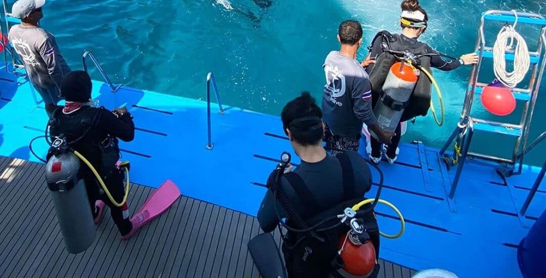 Wetsuited divers with tanks on a boat's blue dive deck, crew helping one at the ladder