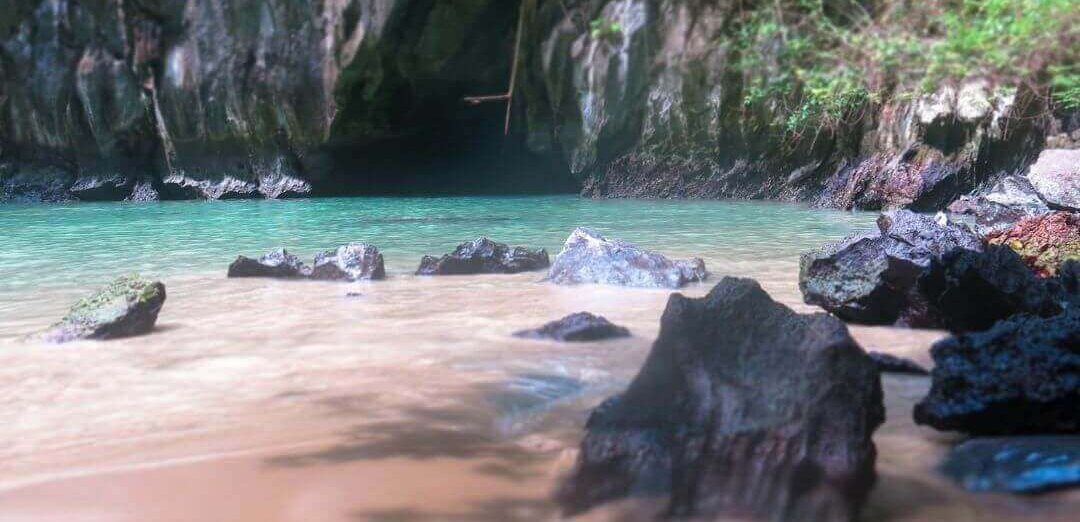 Dark cave mouth in limestone cliffs above a turquoise lagoon and pale sand beach