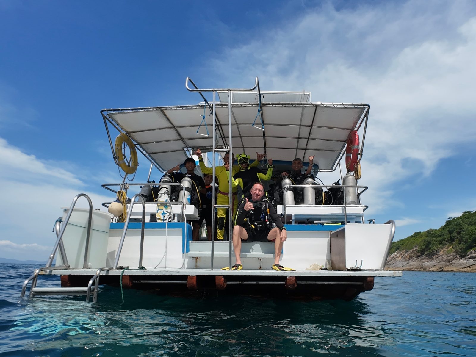 Divers waving from the stern deck of MV Matchanu at anchor off a green island