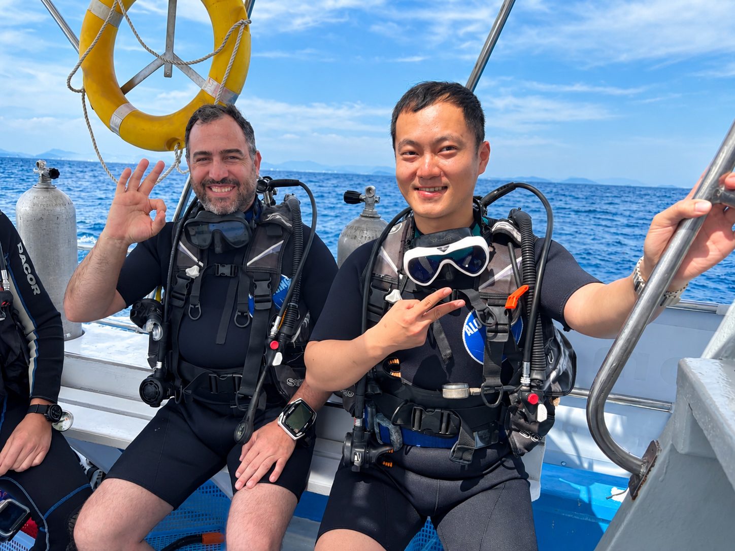 Two divers in their kit on the dive-deck bench, giving the OK sign