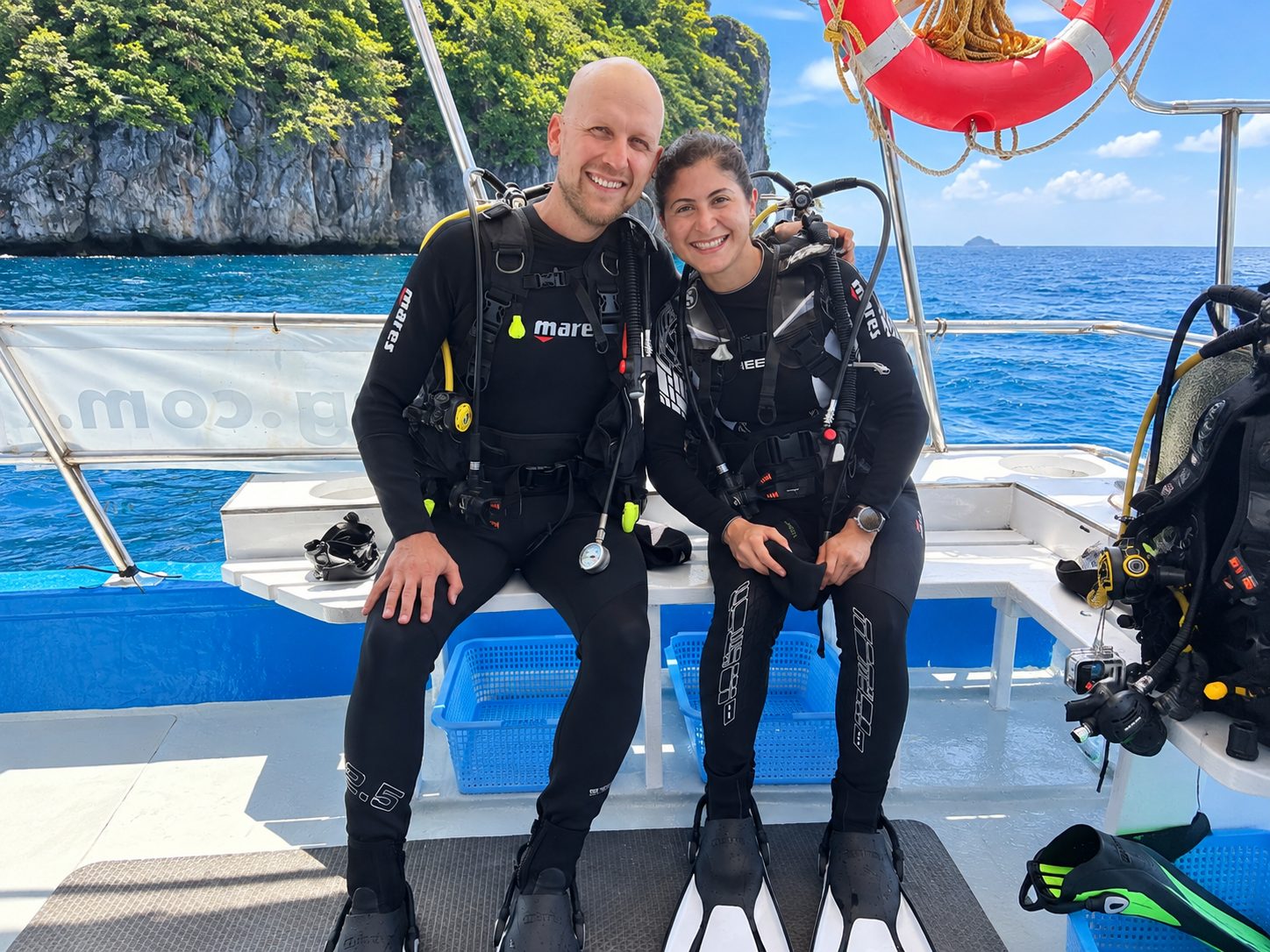 A couple in full kit on the dive-deck bench, a limestone island behind