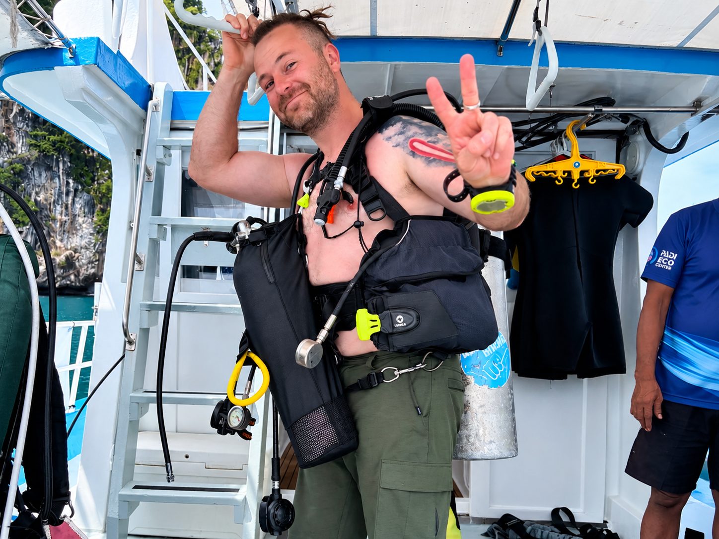 A diver with a pony bottle on his harness, making a peace sign on deck