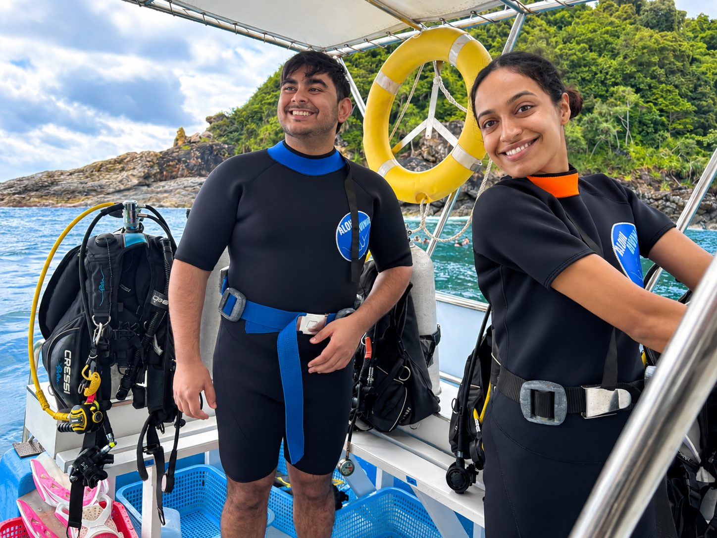 Two guests in wetsuits smiling by the rail, a rocky island behind