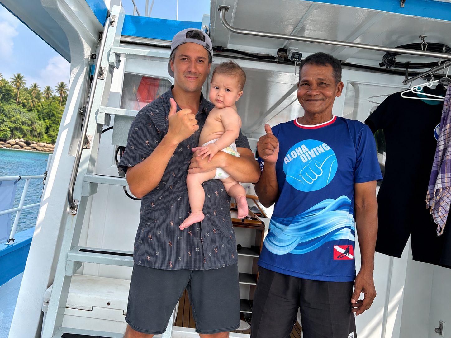 A guest holding his baby on deck beside one of the crew, both giving a thumbs-up