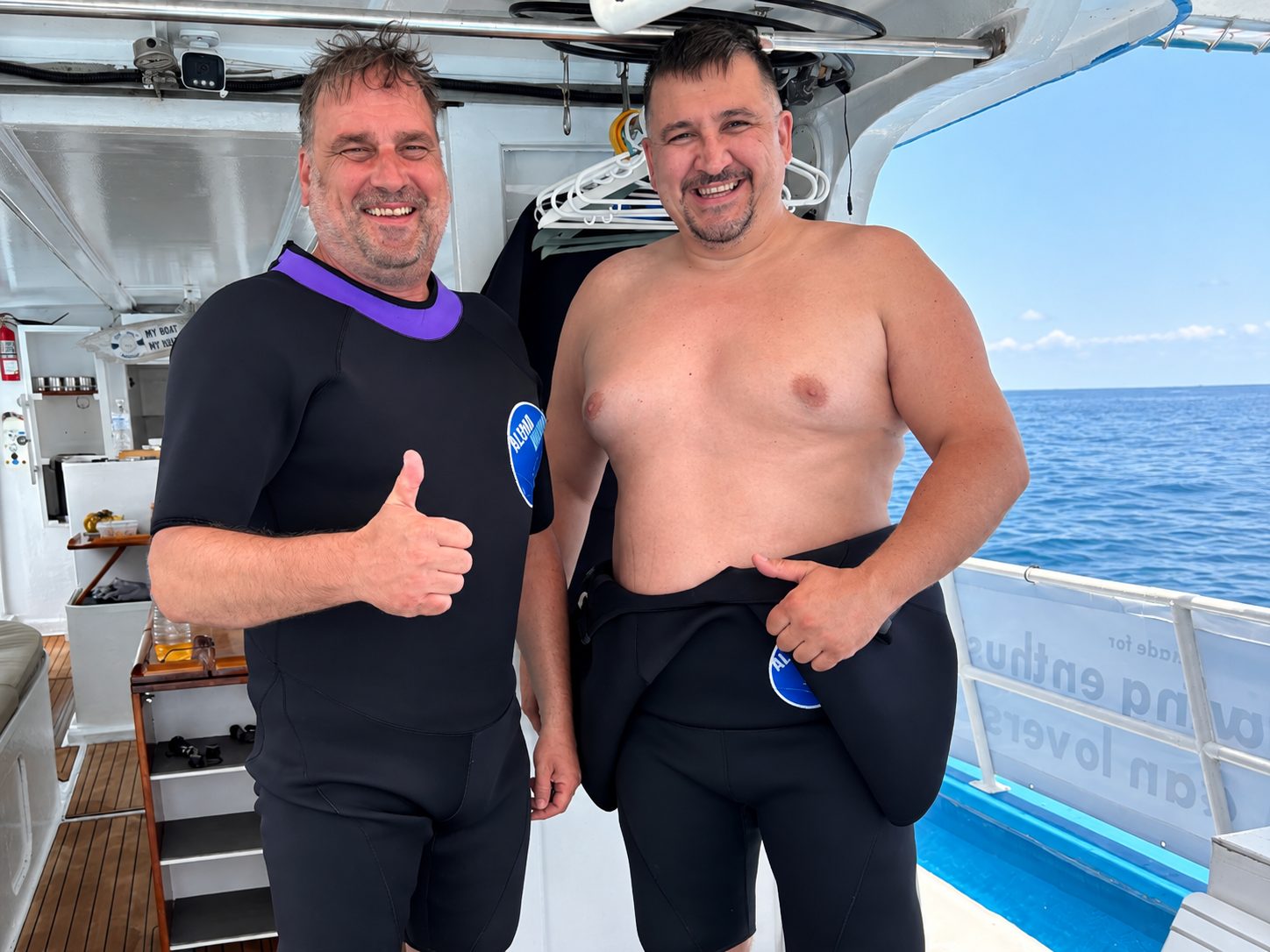 Two guests pulling on their wetsuits on deck, one giving a thumbs-up
