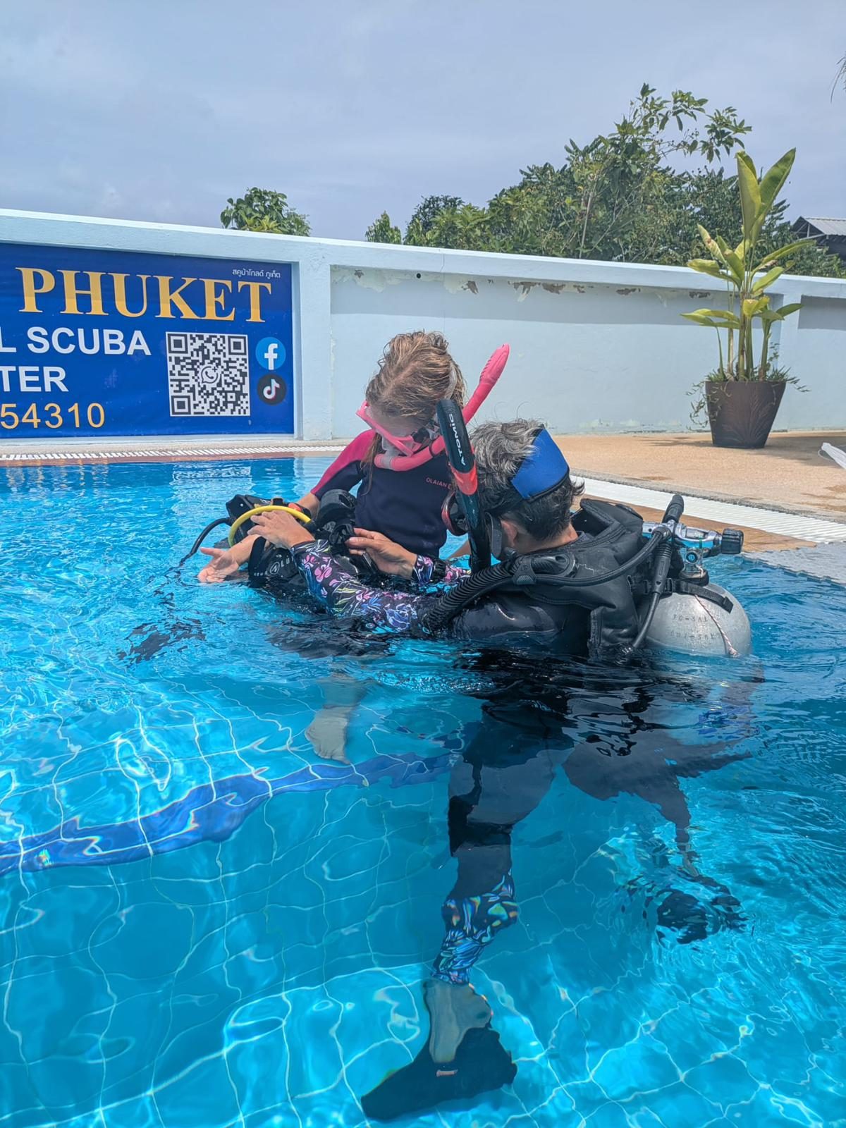 An instructor with a child in scuba gear in the shallow end of the pool, the dive centre sign behind them