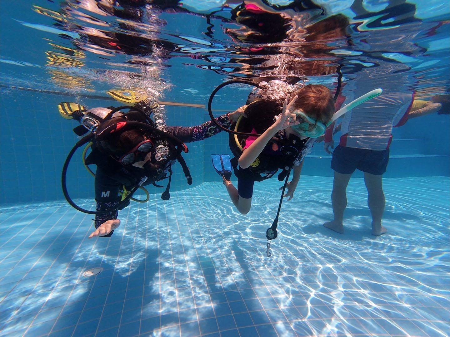 Two children in scuba gear underwater in the training pool, sunlight rippling above them
