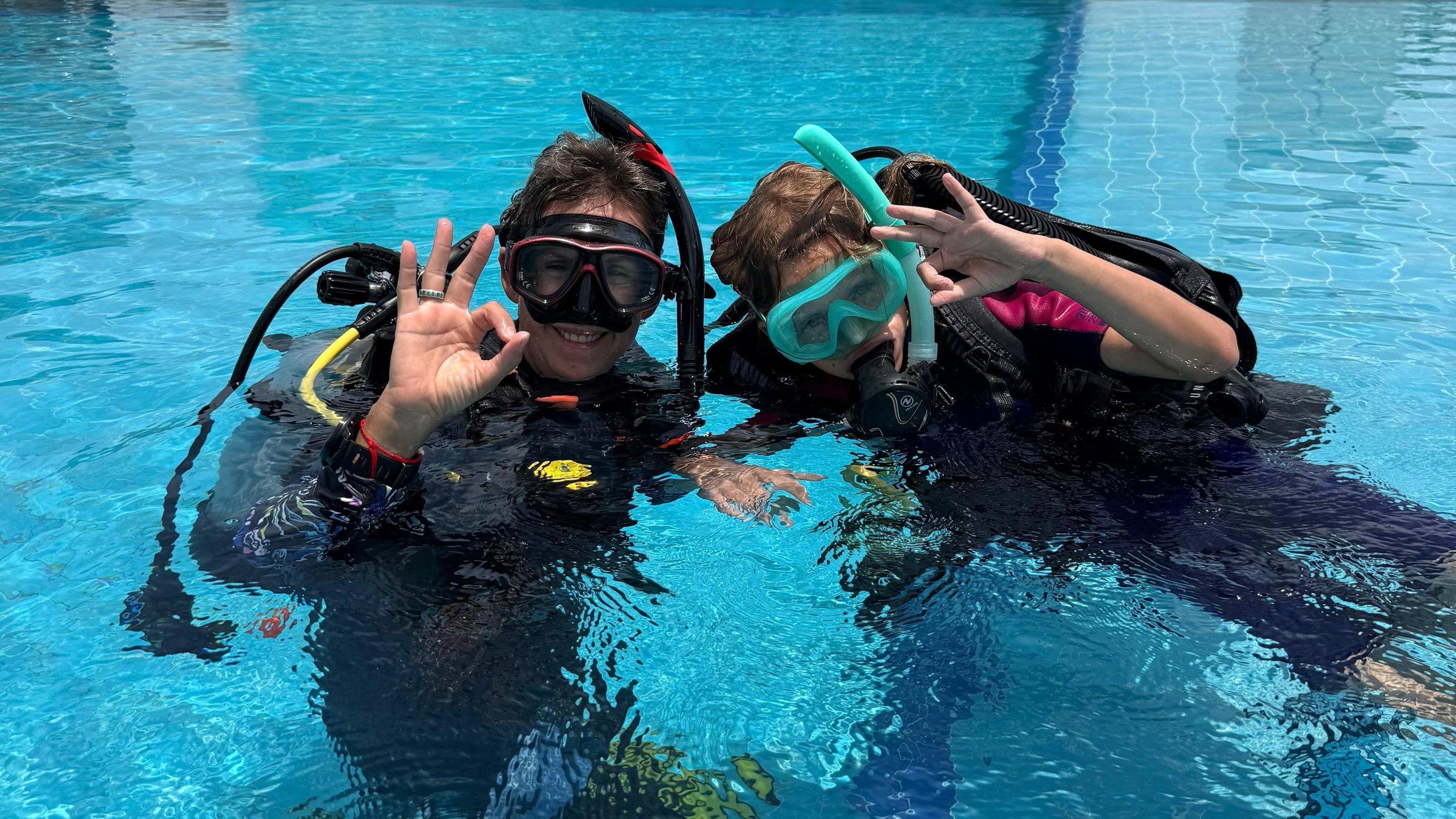 An instructor and a child in scuba gear at the surface of the training pool, both giving the OK sign