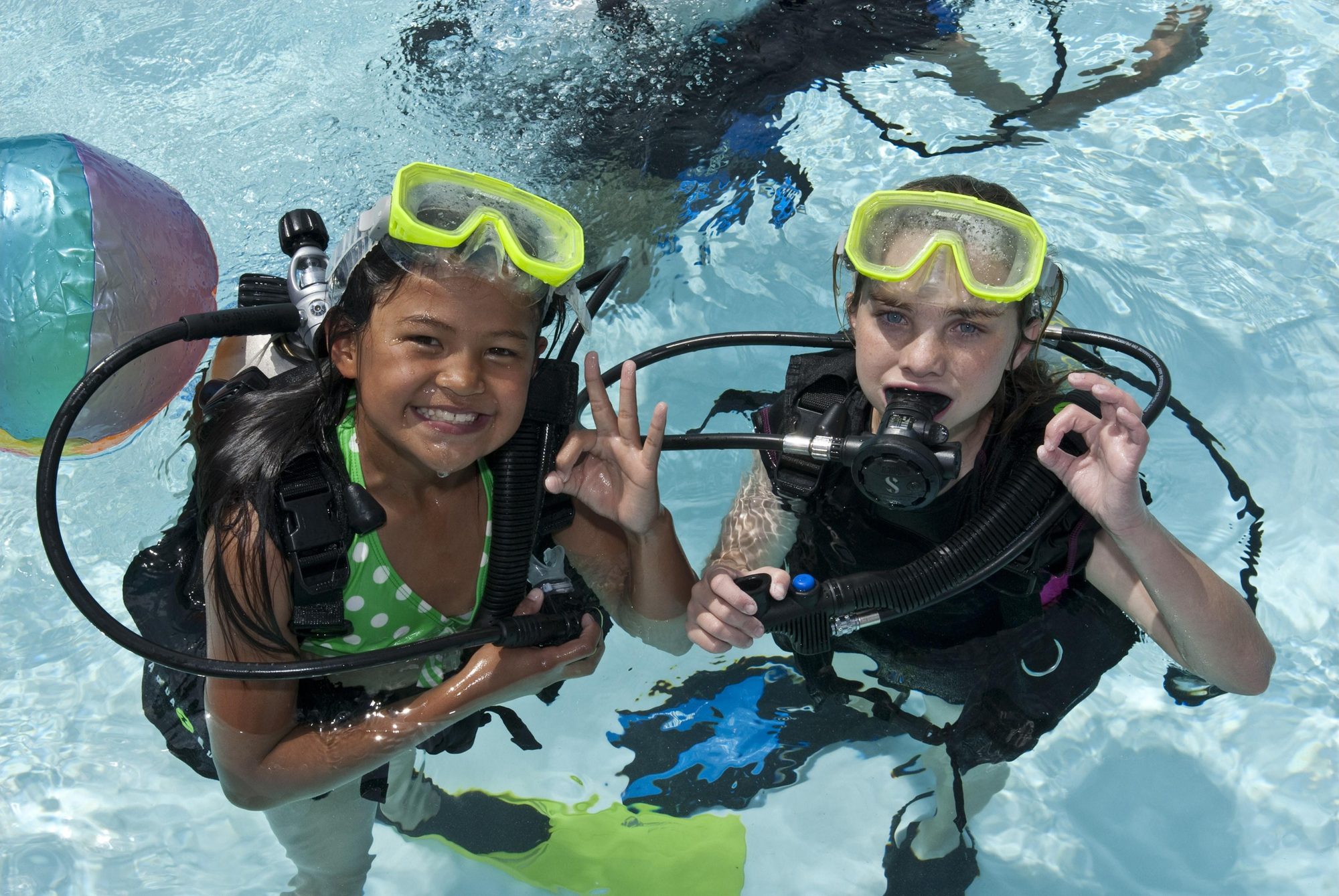 Two children in masks and scuba gear at the surface of a pool, both giving the OK sign