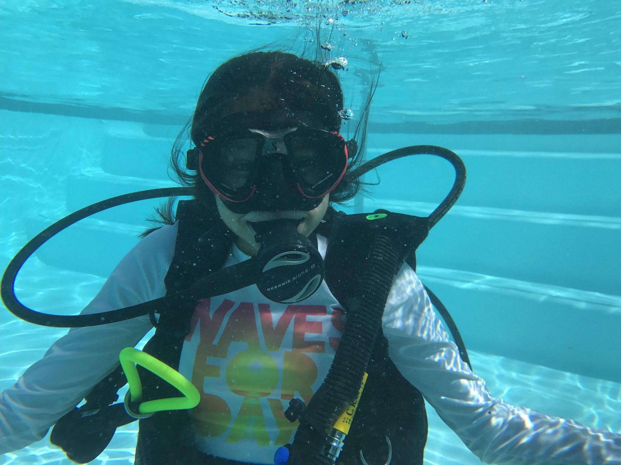 A child in a PADI rash guard underwater in the pool, regulator in, looking at the camera