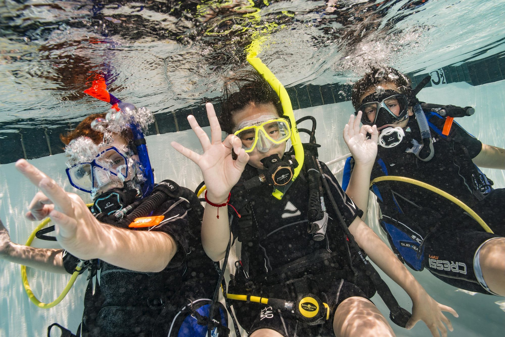 Three young divers at the surface of a pool giving OK signs, a yellow snorkel between them