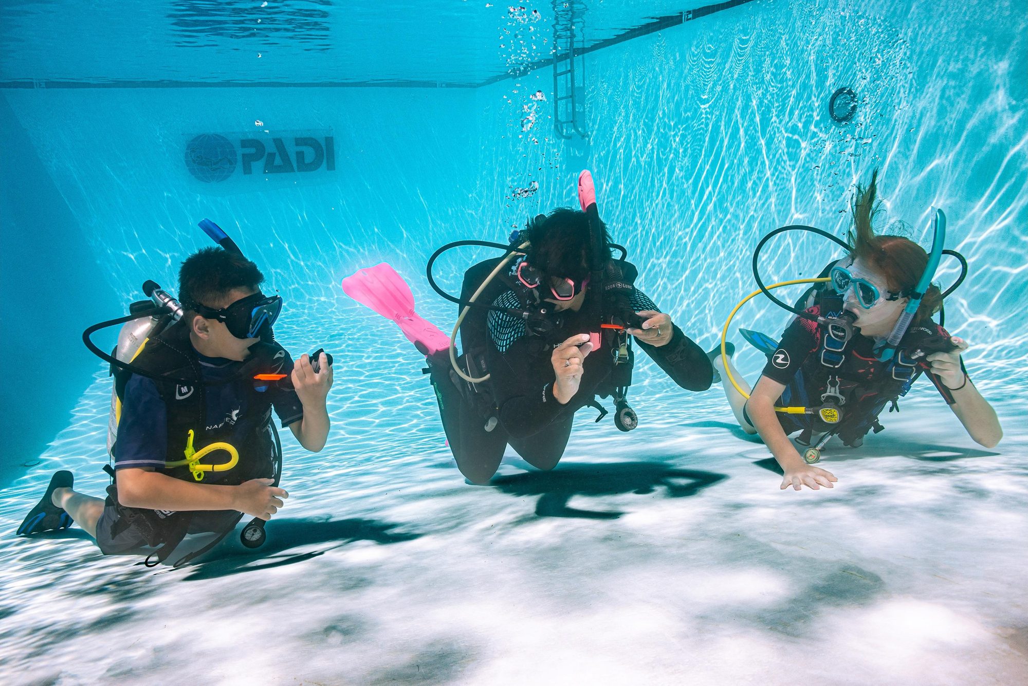 Three young divers kneeling on the pool floor under a PADI banner
