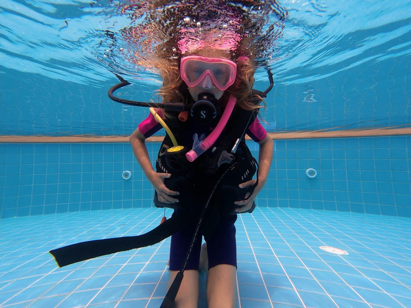 A child in scuba gear underwater in the training pool, light rippling from the surface