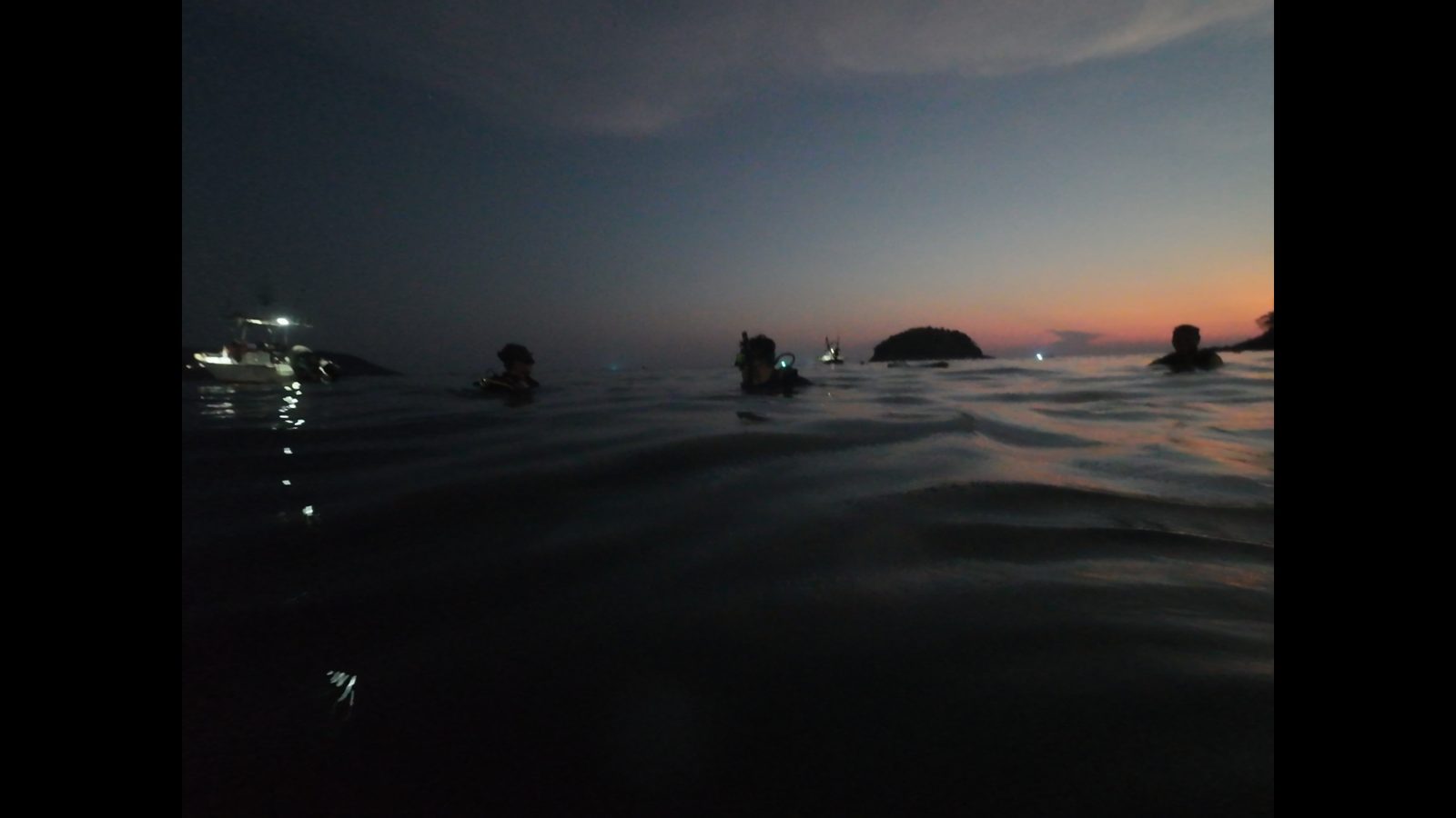 Divers wading into the sea at dusk, the last light on the horizon and boat lights in the bay