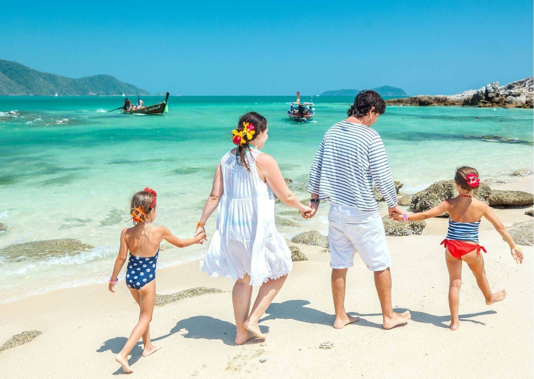 A family of four walking hand in hand along a beach at the water's edge, longtail boats in the bay behind