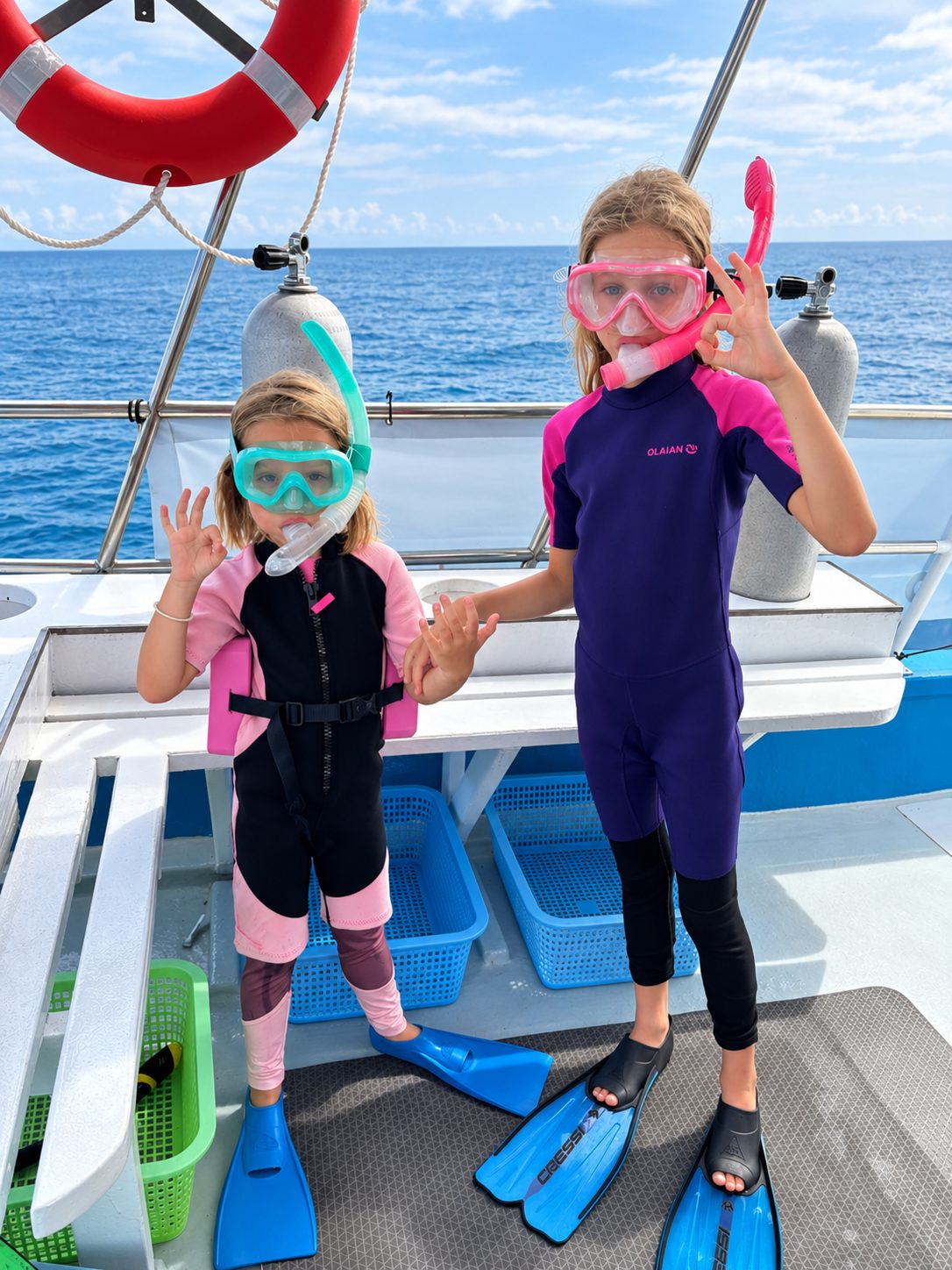 Two children in wetsuits, masks and fins on the deck of a dive boat, waving