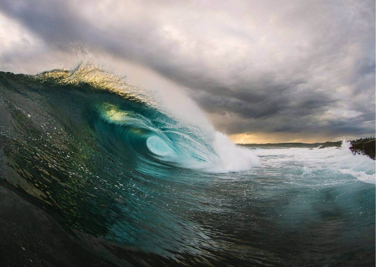 Large wave barrelling at water level beneath heavy grey cloud, low coastline behind