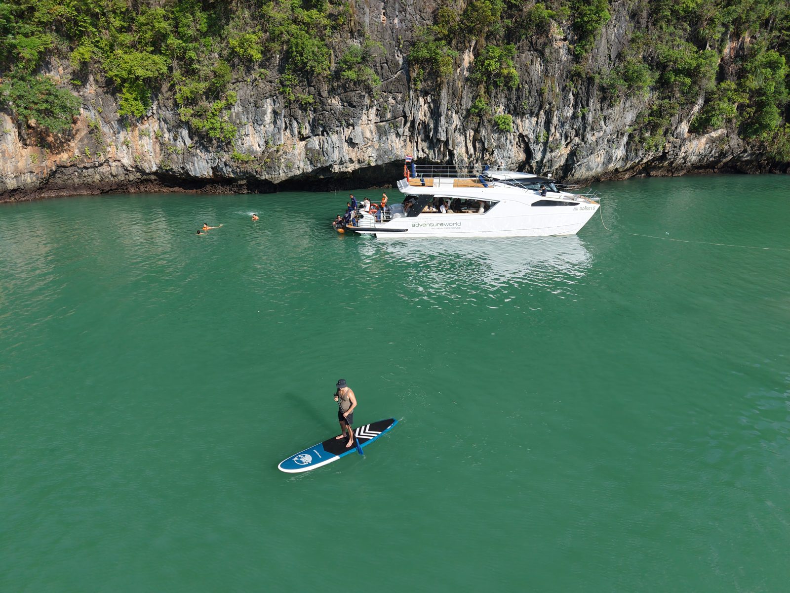 A person on a stand-up paddleboard beside a catamaran in green water below a cliff