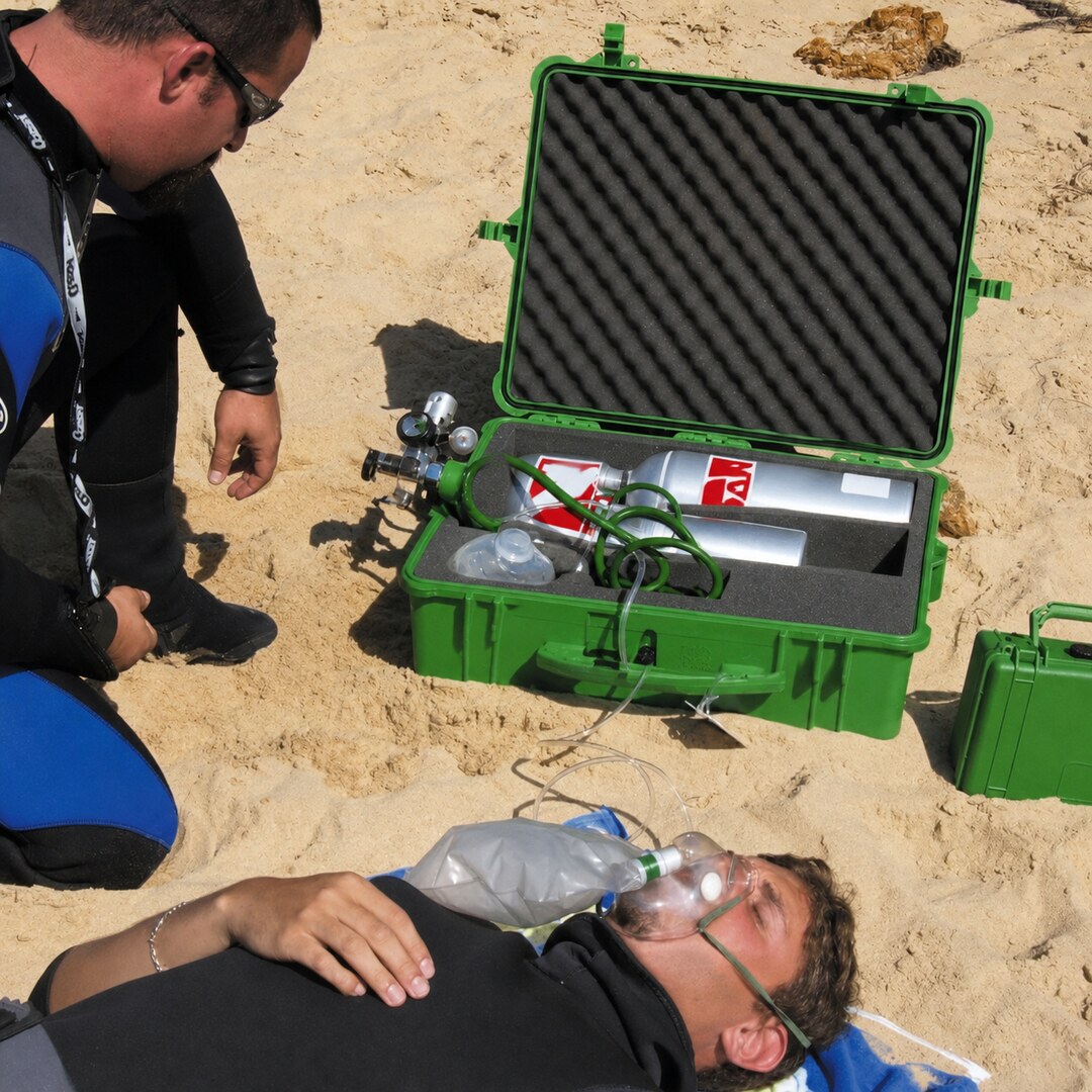Rescuer kneeling beside a casualty on sand with emergency oxygen equipment in an open green case