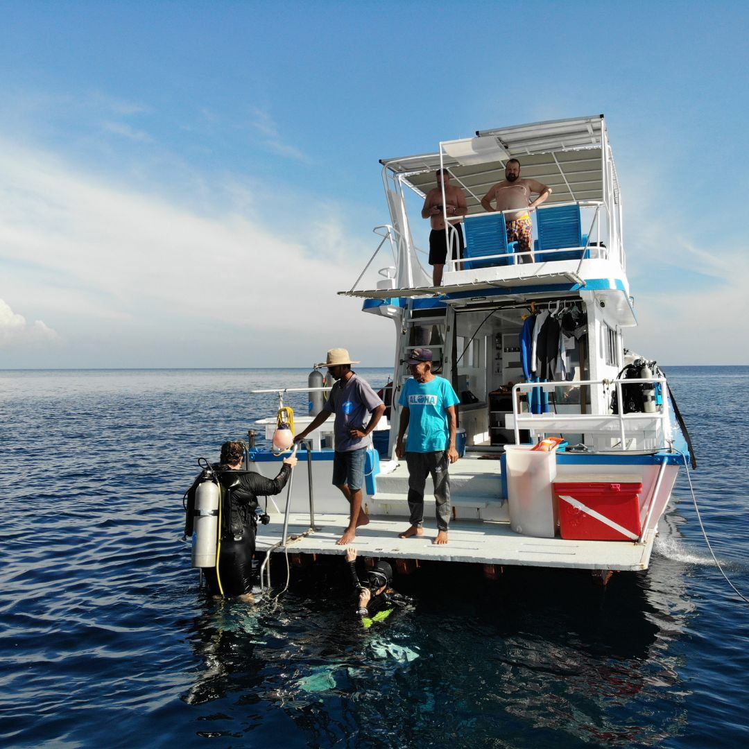 Two divers in the water at a moored dive boat's stern, crew member in an Aloha shirt
