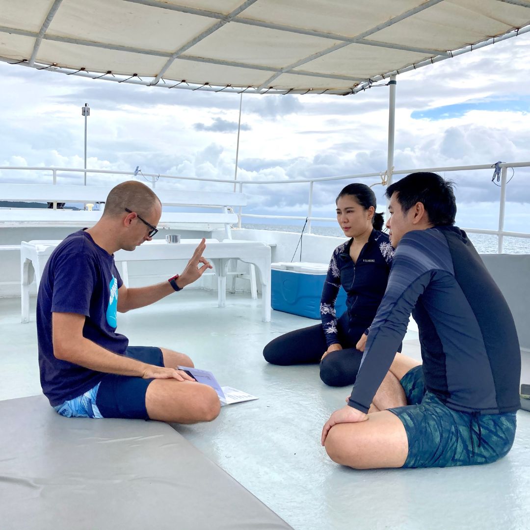 Briefing on a shaded boat deck: a man demonstrates the OK hand signal to two listeners